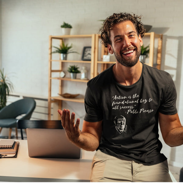 Action is the Foundational Key to Success Pablo Picasso Black T-Shirt man with curly hair laughing with open arms sitting on top of home desk